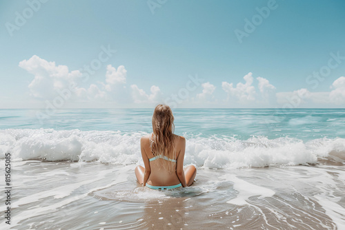 woman in a bikini sits on the shoreline facing the ocean  waves gently crashing around her