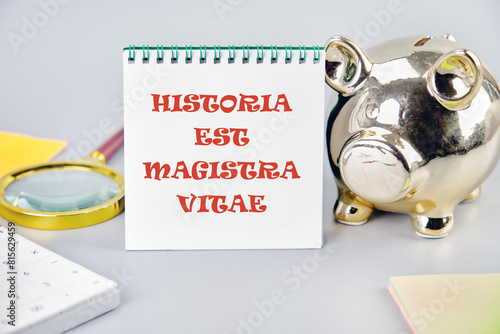 Historia est vitae magistra (History is the tutor of life) Latin phrase on a vertically standing notebook near a piggy bank, a magnifying glass and a calculator photo