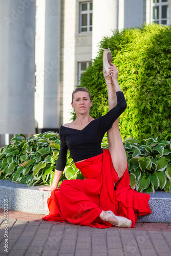 Flexible Winsome Professional Caucasian Ballet Dancer in Red Skirt in Leg Stretching Dance Pose Sitting With Lifted Leg Against Pillars Background.