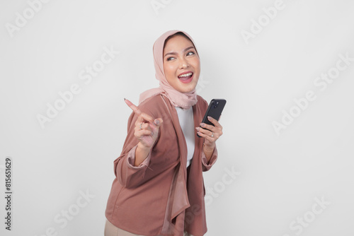 Canvas Print Asian Muslim woman in hijab holding a smart phone while pointing up to the copy space behind over isolated white background