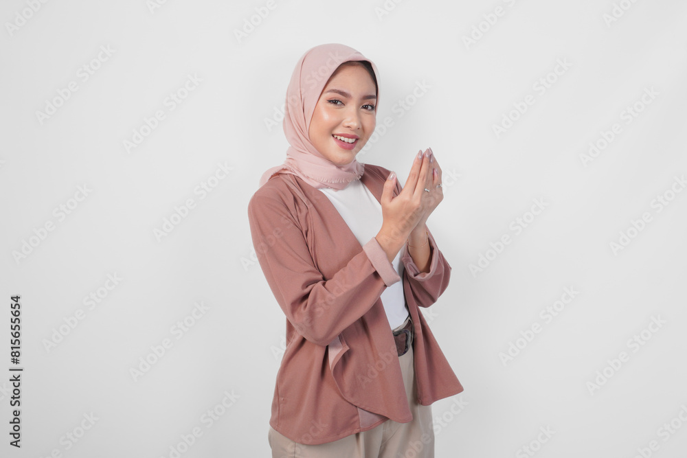 Fototapeta premium Religious young Asian Muslim woman in hijab doing praying gesture while smiling to the camera isolated white background.