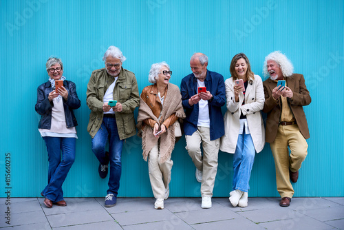 Obraz na płótnie Group of elderly Caucasian laughing together using cell phone standing leaning on blue wall