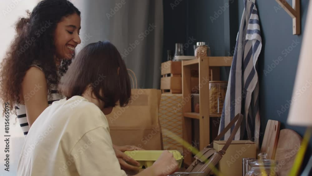 Medium shot of Caucasian girl with cerebral palsy and her Biracial female friend chatting while unpacking paper bags on kitchen table after doing grocery shopping together Medium shot of Caucasian girl with cerebral palsy and her Biracial female friend chatting while unpacking paper bags on kitchen table after doing grocery shopping together