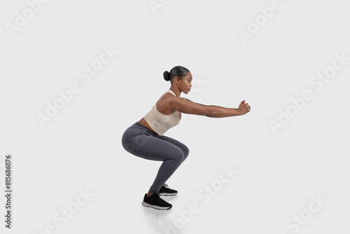 African American woman is seen on a white background, demonstrating a squat exercise. Her body is positioned with feet shoulder-width apart, side view