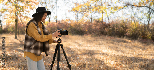 beautiful Caucasian woman doing a photo shoot in the autumn forest. A professional photographer photographs a landscape on a sunny day using a camera and a tripod. banner