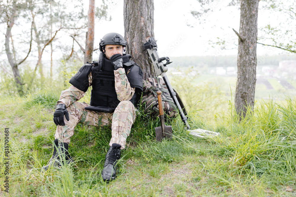 A man in a military uniform and a bulletproof vest sits in the forest near a metal detector and a military backpack. A man pauses in the work of demining the territory A man in a military uniform and a bulletproof vest sits in the forest near a metal detector and a military backpack. A man pauses in the work of demining the territory
