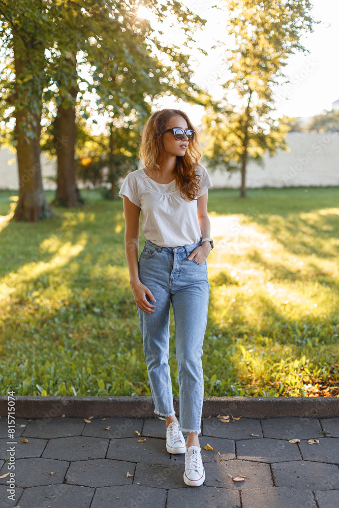 Beauty pretty woman with red hair with sunglasses in summer clothes with t-shirt and jeans poses in the park Beauty pretty woman with red hair with sunglasses in summer clothes with t-shirt and jeans poses in the park
