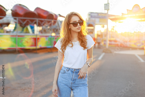 Summer beautiful stylish girl with red hair with fashionable sunglasses in a white vintage T-shirt walks on the street at sunset