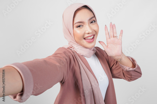 Cheerful young Asian Muslim woman in hijab posing for a selfie with waving hand hello isolated over white background.