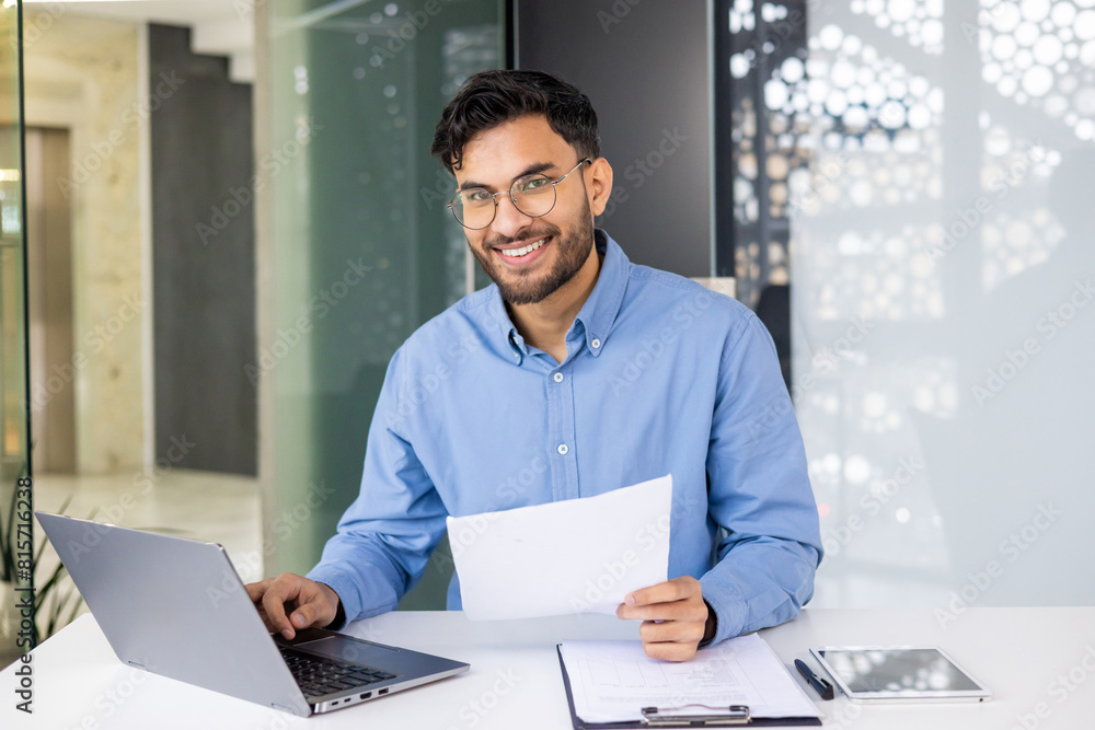 Smiling businessman working on laptop and reviewing documents at office desk Smiling businessman working on laptop and reviewing documents at office desk