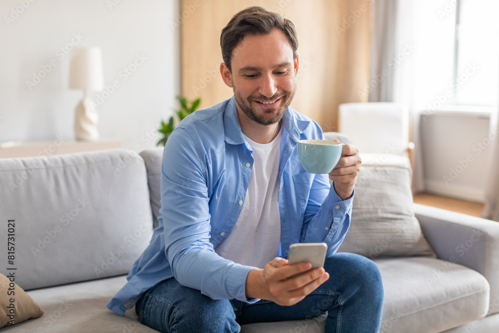 A man is sitting on a couch, holding a cup of coffee in one hand and looking at his phone in the other. He appears relaxed and focused on his phone screen. A man is sitting on a couch, holding a cup of coffee in one hand and looking at his phone in the other. He appears relaxed and focused on his phone screen.