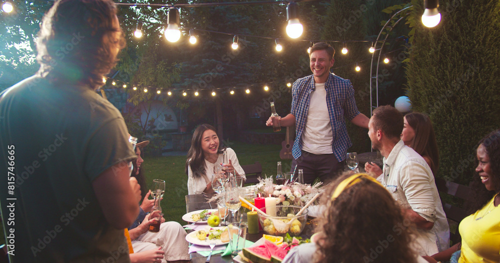 Happy multi-ethnic people having fun and playing at garden party in evening. Caucasian boys and girls drinking and talking. African American and Asian females laughing. Party games. Fun concept Happy multi-ethnic people having fun and playing at garden party in evening. Caucasian boys and girls drinking and talking. African American and Asian females laughing. Party games. Fun concept