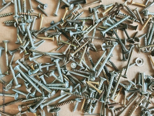 Assorted Screws in Different Sizes and Colors on Light Brown Background. Close-Up View of Various Screws Scattered in Random Pattern. Industrial Hardware Concept for Construction and Repair Projects