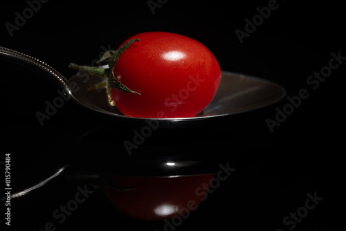Close-up of a small red tomato lying on a metal spoon. The spoon and the tomato are reflected against a dark background