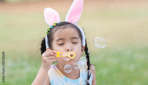 Happy 6 year old Asian little girl with bunny ears blowing soap bubbles in park, having fun, sunny day. Portrait of cute child playing outdoors in summer in nature