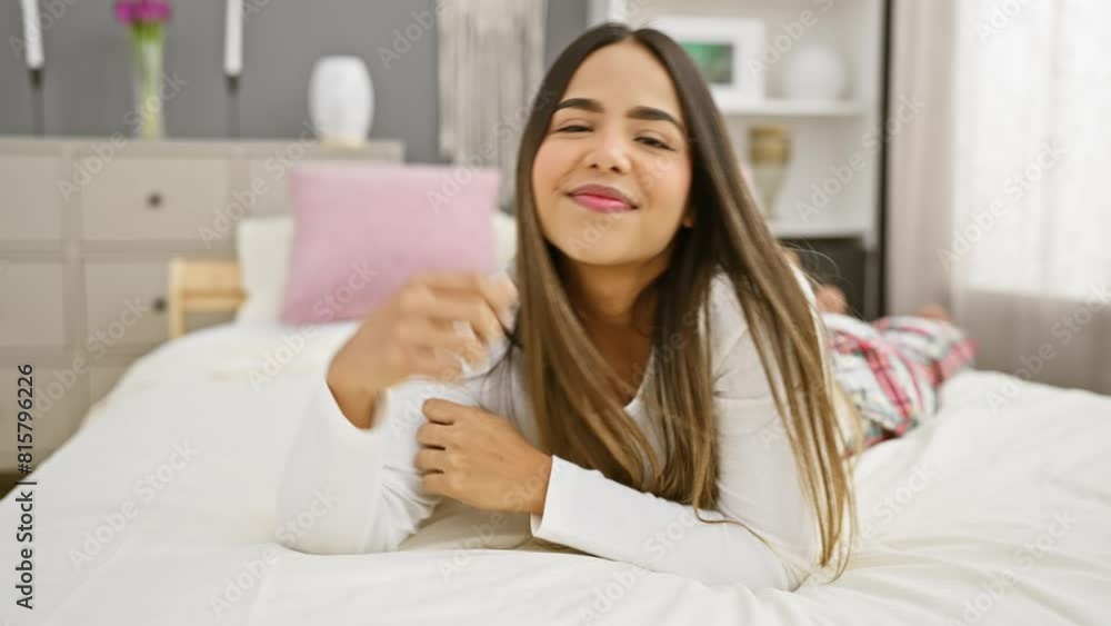 A young hispanic woman smiles while lying on a white bed in a well-lit modern bedroom, embodying relaxation and comfort. A young hispanic woman smiles while lying on a white bed in a well-lit modern bedroom, embodying relaxation and comfort.