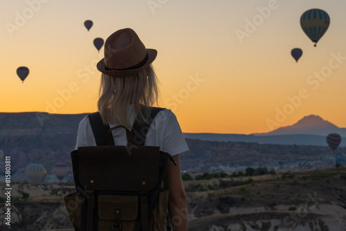 Young tourist in a hat with a backpack enjoys the view of balloons and the beginning of sunrise in Cappadocia, Turkey. Beautiful woman on top of a mountain enjoys a beautiful view.