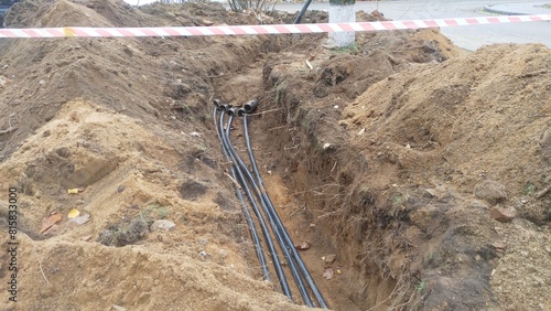 A trench was dug next to trees and the asphalt road to replace cables, pipes and other utilities. The excavated soil lies on the sides of the trench and is taped for safety. Autumn and fallen leaves