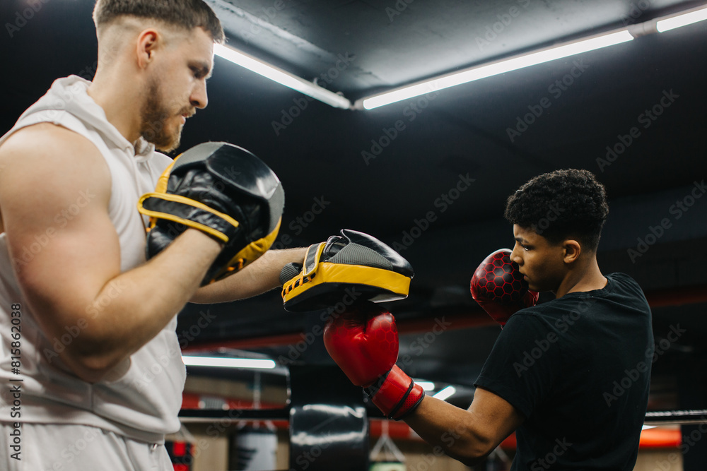 A male trainer helps a teenage boxer with striking techniques in a boxing ring. A male trainer helps a teenage boxer with striking techniques in a boxing ring.