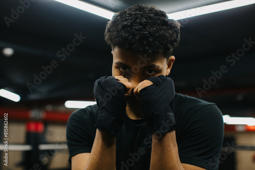 A determined young boxer is looking at the camera while standing in a fighting stance with boxing bandages on his fists. Sporty young man training in boxing gym.