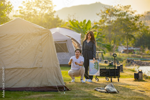 Young tourist asian woman on vacation holding map and looking young man setting tent in camping ground near river with camping tent in the background. Beautiful view. 