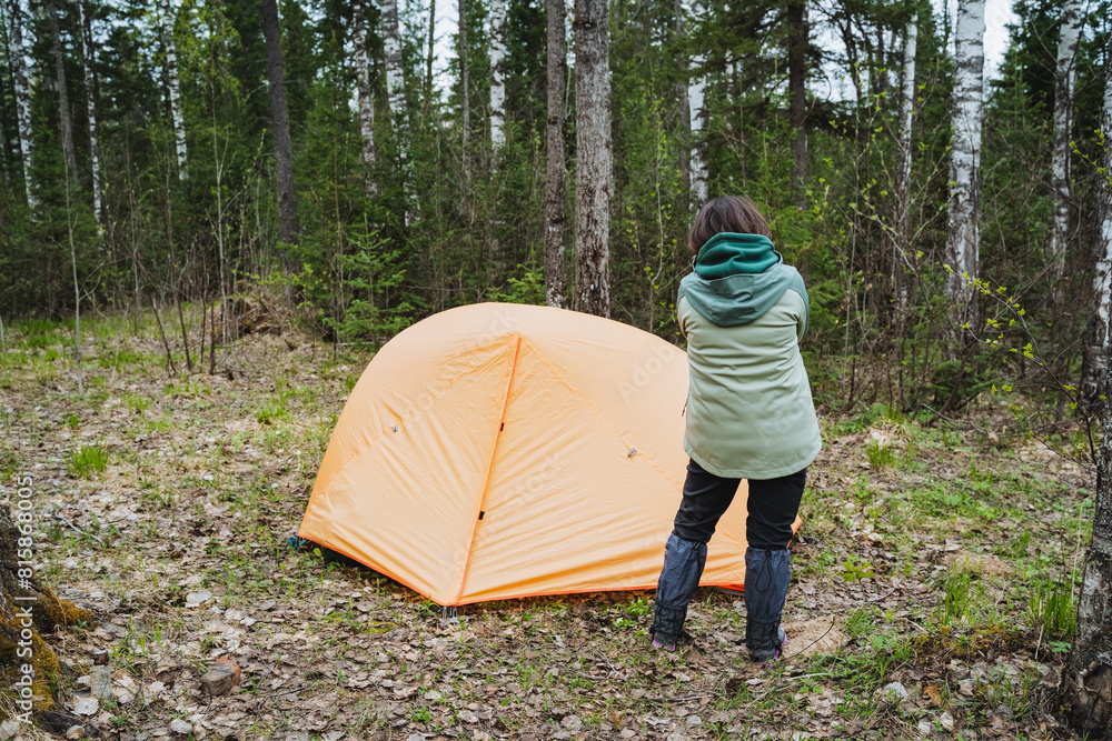 A woman stands by orange tent in woodland with grass and trees