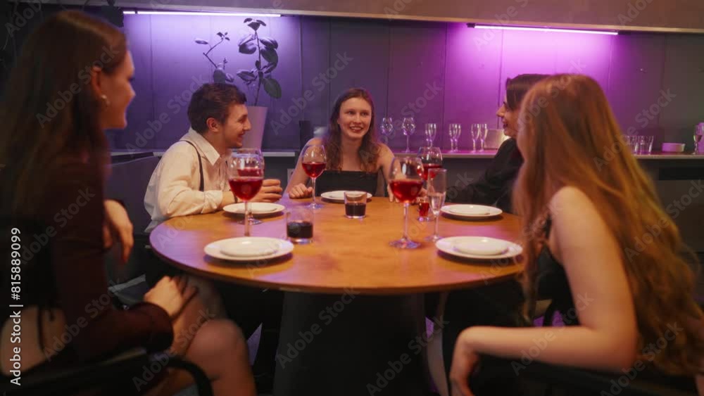 Group of friends talking in night bar. Young cheerful girl emotionally tells the story to friends laughing drinking alcohol cocktails, sitting at table, having fun together. Party friendship concept. Group of friends talking in night bar. Young cheerful girl emotionally tells the story to friends laughing drinking alcohol cocktails, sitting at table, having fun together. Party friendship concept.