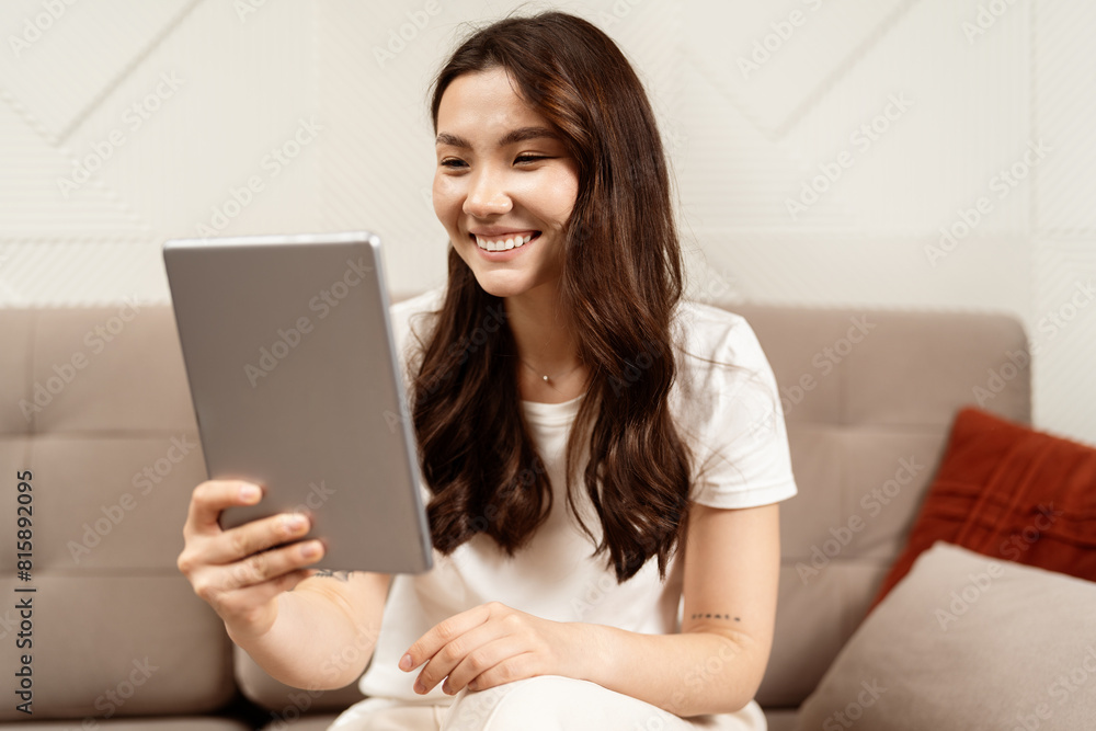 Young Woman Using Tablet At Home: A cheerful young woman enjoys using a digital tablet, sitting comfortably on a couch, portraying technology and lifestyle.