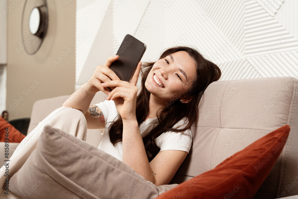 Young Woman Enjoying Social Media On Smartphone At Home, Lying Comfortably On Couch With Happy Expression, Modern Living Room Interior Young Woman Enjoying Social Media On Smartphone At Home, Lying Comfortably On Couch With Happy Expression, Modern Living Room Interior
