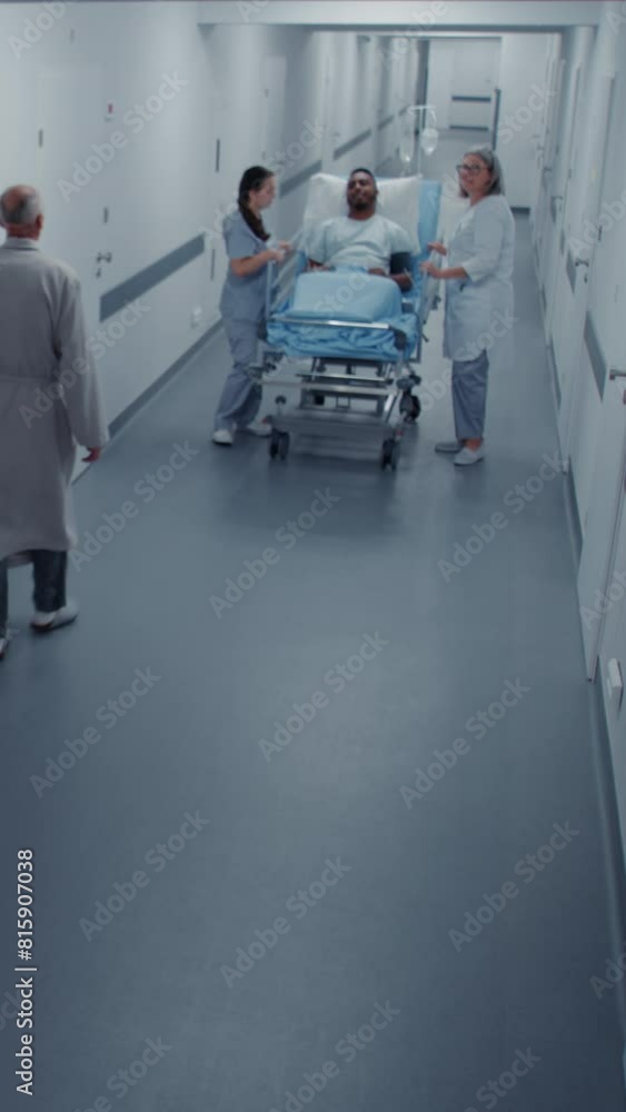 Female doctor and nurse stand in hospital corridor with African American patient on gurney. Professional medics transport man to operating room on surgery. Security camera view. Vertical shot Female doctor and nurse stand in hospital corridor with African American patient on gurney. Professional medics transport man to operating room on surgery. Security camera view. Vertical shot