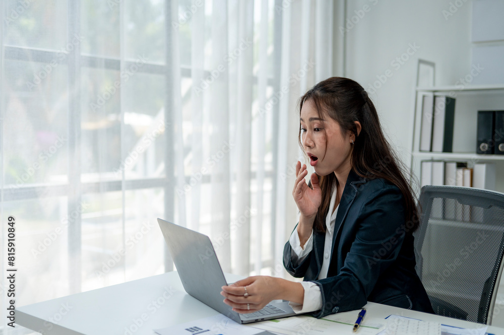 A woman is sitting at a desk with a laptop open in front of her. She is looking at the screen with a shocked expression on her face A woman is sitting at a desk with a laptop open in front of her. She is looking at the screen with a shocked expression on her face