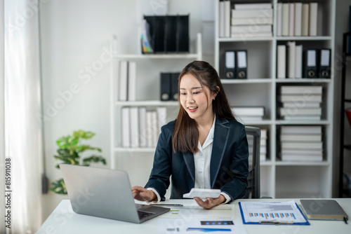 A woman in a business suit is sitting at a desk with a laptop and a calculator. She is smiling and she is enjoying her work