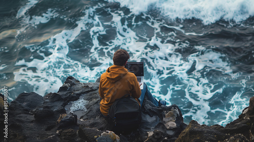 A person in a yellow jacket sits on rocky cliffs using a laptop, overlooking turbulent ocean waves.