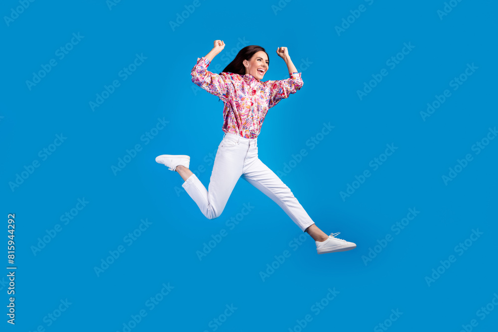 Full length photo of funky cheerful lady dressed print blouse rising fist jumping high running empty space isolated blue color background Full length photo of funky cheerful lady dressed print blouse rising fist jumping high running empty space isolated blue color background
