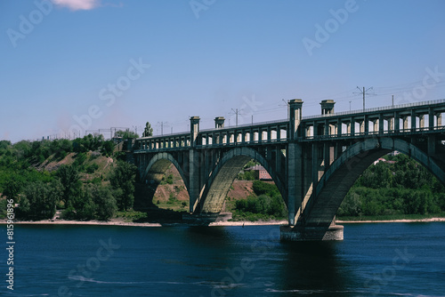 Vintage arched bridge crossing over a river, featuring railway tracks on top and surrounded by lush greenery with a hint of urban buildings in the background © Andrii Chagovets