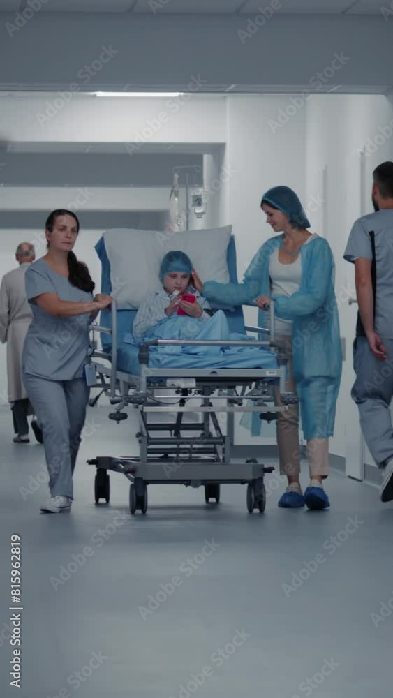 Nurse and mother of child push gurney with young girl in clinic hallway. Female doctor meets lying on stretcher kid and talks with her before operation. Medical personnel work in emergency department. Nurse and mother of child push gurney with young girl in clinic hallway. Female doctor meets lying on stretcher kid and talks with her before operation. Medical personnel work in emergency department.