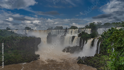 Beautiful tropical waterfall landscape. Cascading streams collapse from ledges into the bed of a stormy river. Splashes  fog. Clouds in the blue sky. Lush green vegetation. Iguazu Falls. Argentina.