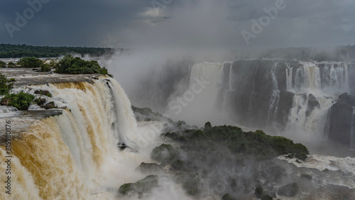 An impressive waterfall landscape. Streams of white foaming water collapse from a ledge in the gorge. Spray and fog rise like a cloud into the sky. Tropical vegetation. Iguazu Falls. Brazil.