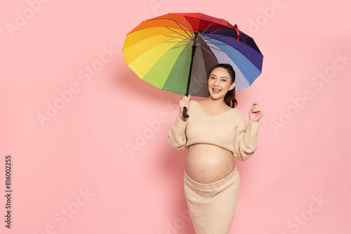 Portrait of Happy Asian pregnant woman holding rainbow umbrella isolated on pink background, In the rainy season with pregnancy concept