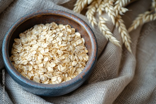 Close-up of raw oat flakes in a rustic ceramic bowl beside wheat ears on a textured linen background