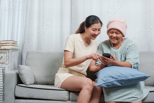 Senior Asian woman in knitted hat reviews phone content, directed by younger woman, both seated on gray sofa. Older lady in blue-green tunic and pink toque concentrates on smartphone