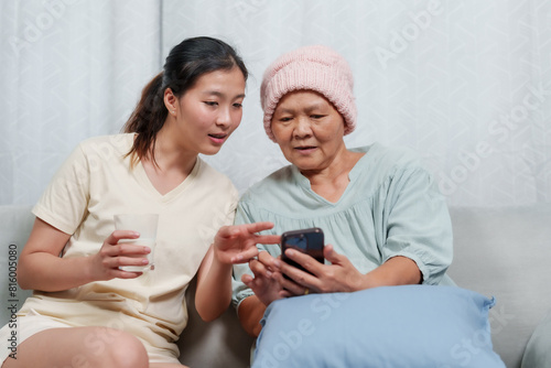 Young woman holding glass of milk showing elderly woman something on smartphone. Elderly woman wearing pink beanie, indicating cancer treatment. Atmosphere is warm, caring. sharing moment, support.
