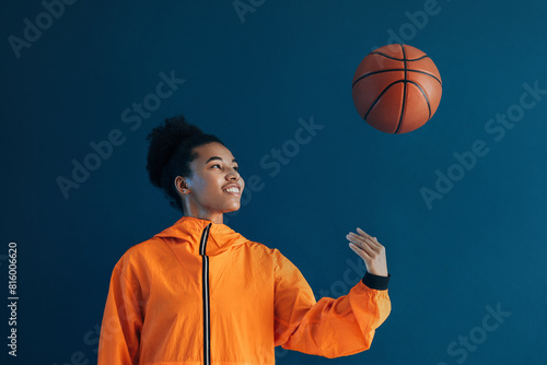 Female basketball player tossing a basketball over a blue background. Young woman in orange sportswear tossing basketball up in studio.