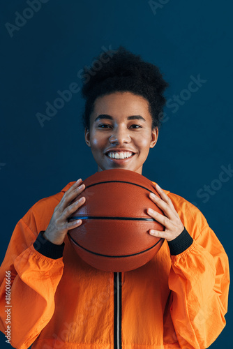 Portrait of a young smiling basketball player. Cheerful female in orange sportswear holding basketball over blue backdrop.