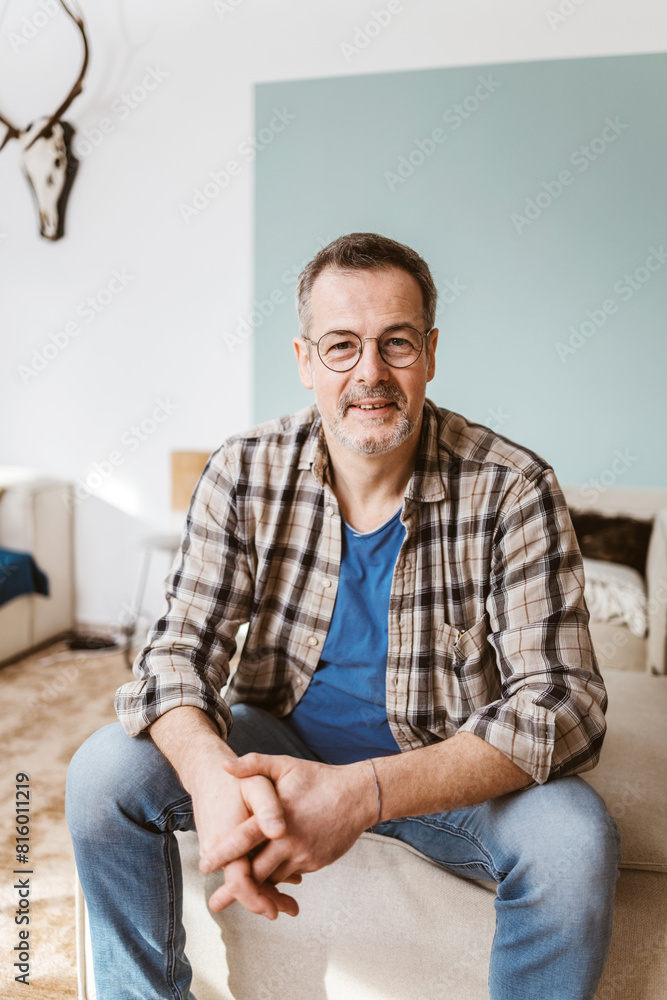 Casual mature man wearing glasses and plaid shirt, sitting indoors with a relaxed expression. Comfortable and confident ambiance.
