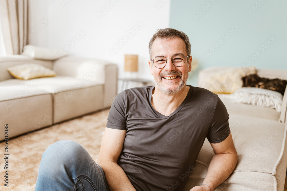 A cheerful middle-aged man with glasses relaxing on the floor of his cozy living room, showing comfort and happiness. A cheerful middle-aged man with glasses relaxing on the floor of his cozy living room, showing comfort and happiness.