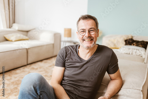 A cheerful middle-aged man with glasses relaxing on the floor of his cozy living room, showing comfort and happiness.