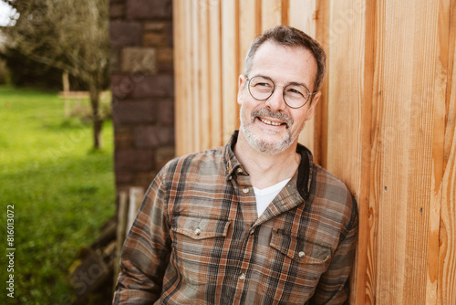 A smiling mature man wearing a plaid shirt is standing outdoors, leaning against a wooden wall on a sunny day.