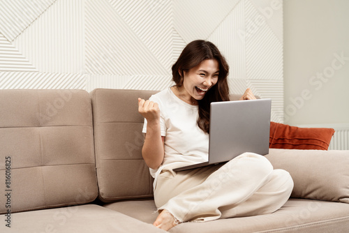 Joyful Woman Celebrating Success On Laptop In Modern Living Room, Asian Female Excited By Good News Online, Technology Win Concept