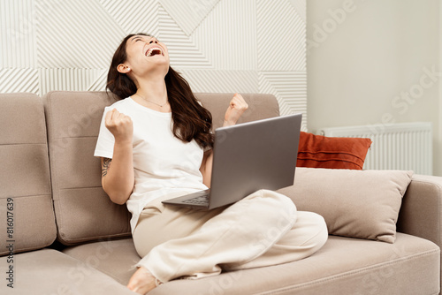 Happy Woman Celebrating Success At Home With Laptop. Joyful Female On Sofa Feeling Excited, Achievement Concept, Modern Living Room.